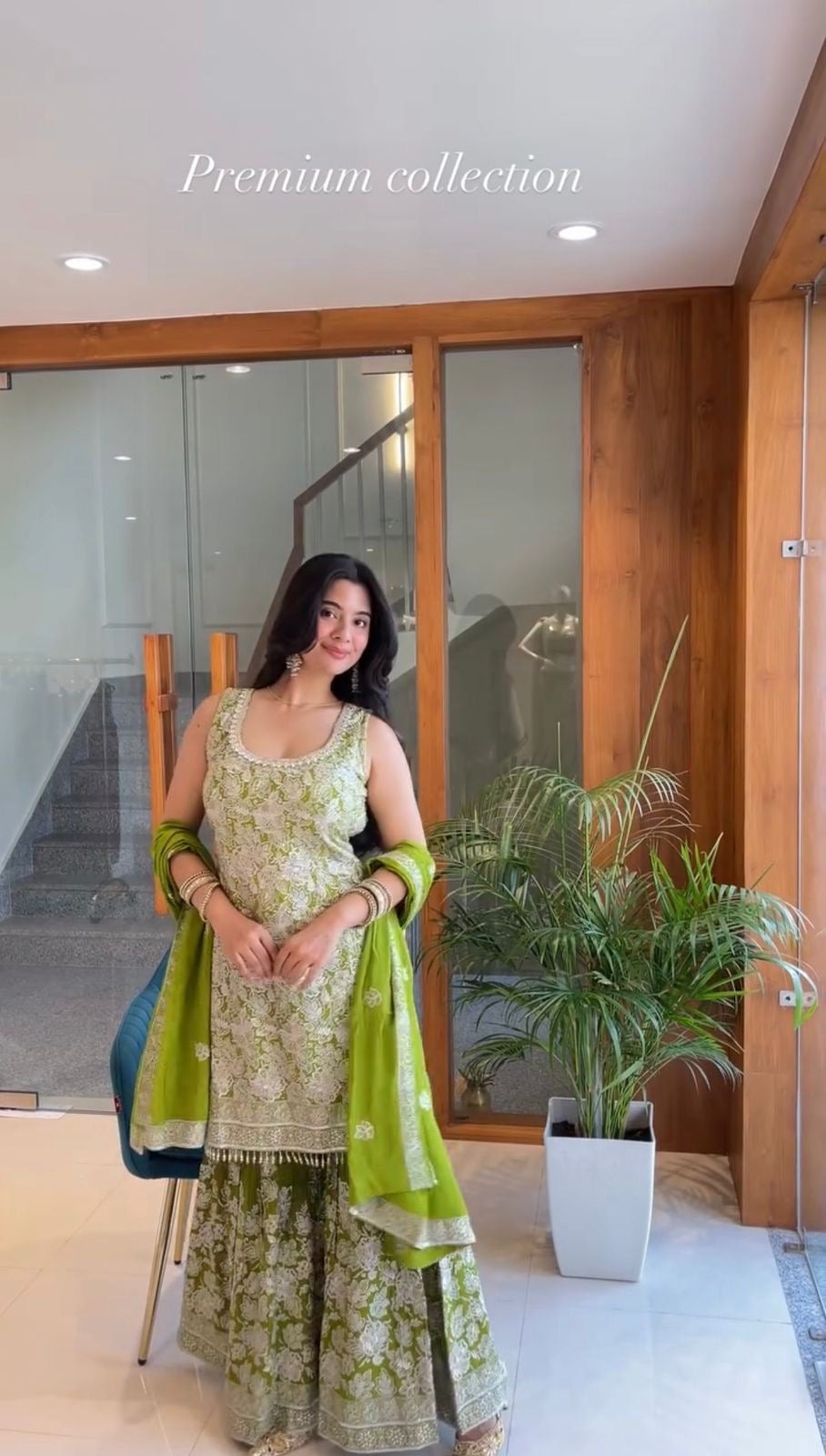 Woman in a green and white traditional outfit standing indoors with plants and wooden panels in the background.