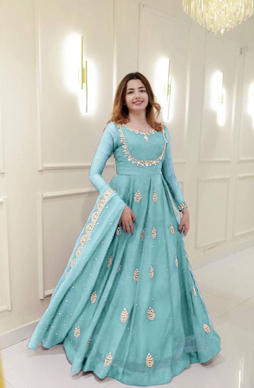 Woman wearing a light blue traditional dress with gold embroidery in a white room.