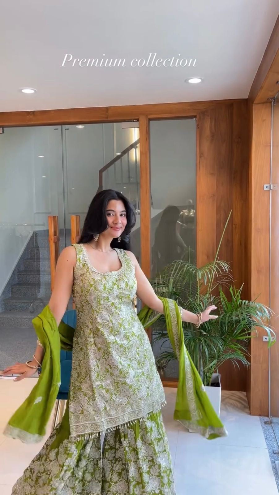 Woman in a green and white traditional outfit standing indoors with plants and a staircase in the background.