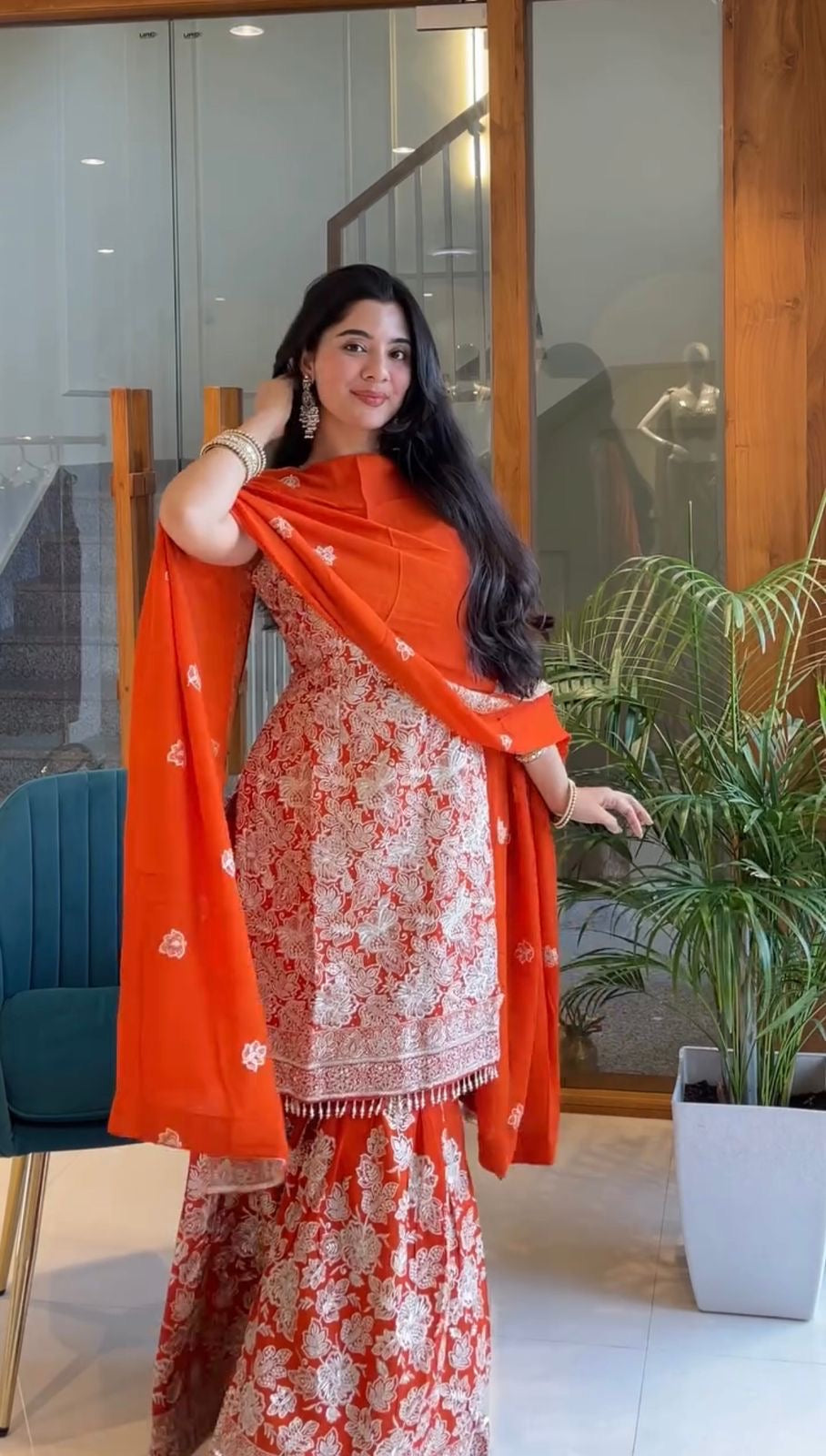 Woman in an orange and white traditional outfit standing indoors with a plant in the background