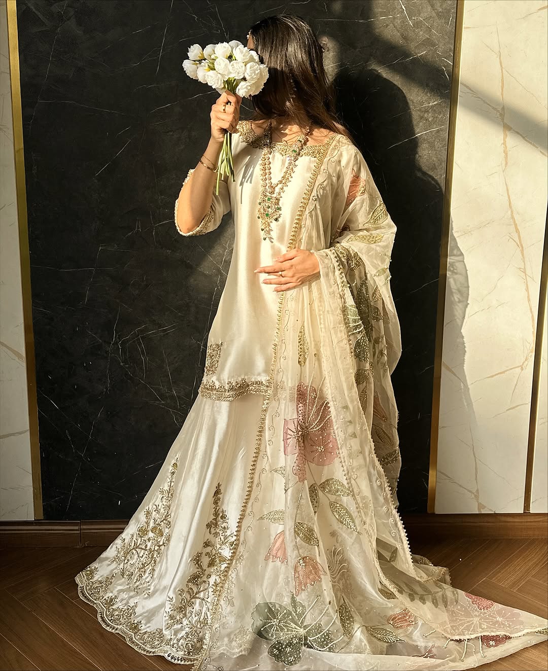 Woman in a white embroidered dress holding flowers against a marble wall.