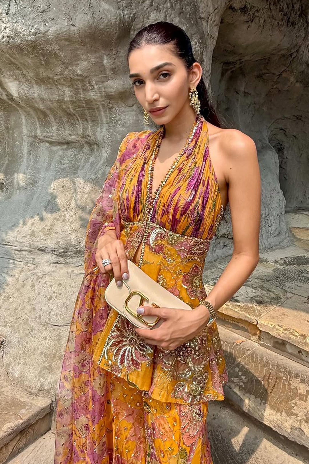 Woman in a colorful dress holding a beige handbag against a stone wall background