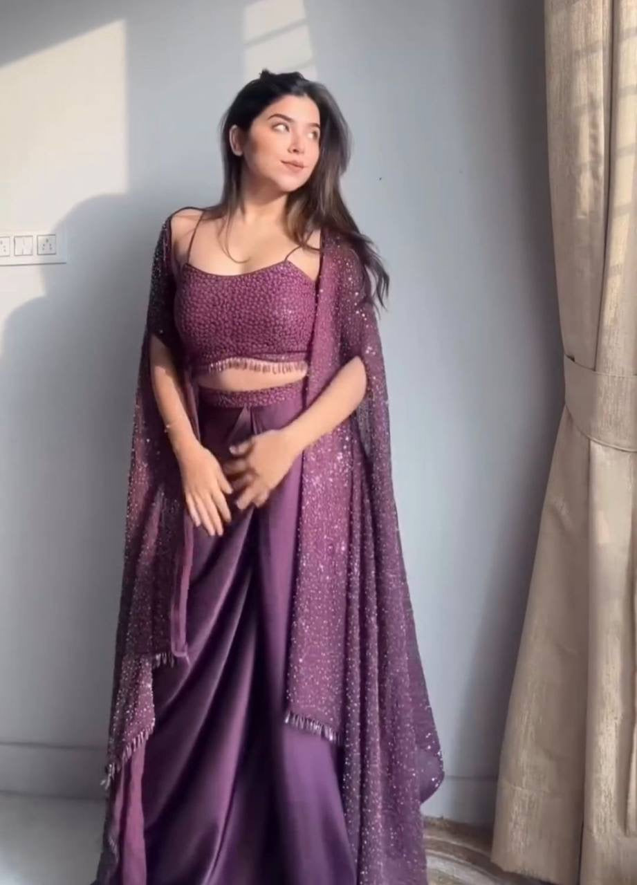 Woman in a purple saree with a matching blouse standing against a light gray wall.