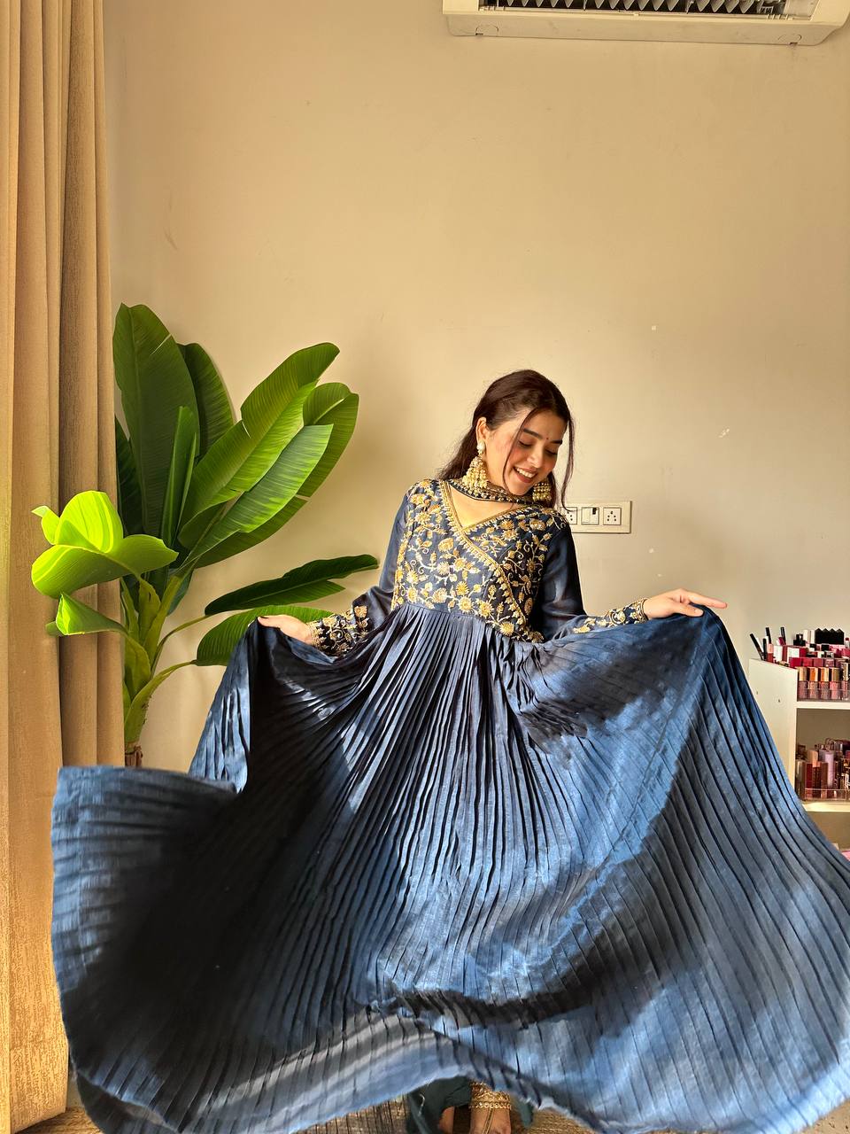 Woman holding a long, pleated blue dress in a room with a plant and wall socket.