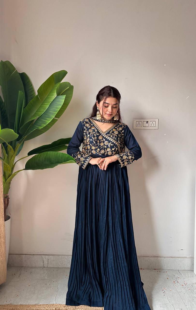 Woman wearing a traditional outfit with a floral top and pleated skirt, standing against a plain wall with a plant to the side.