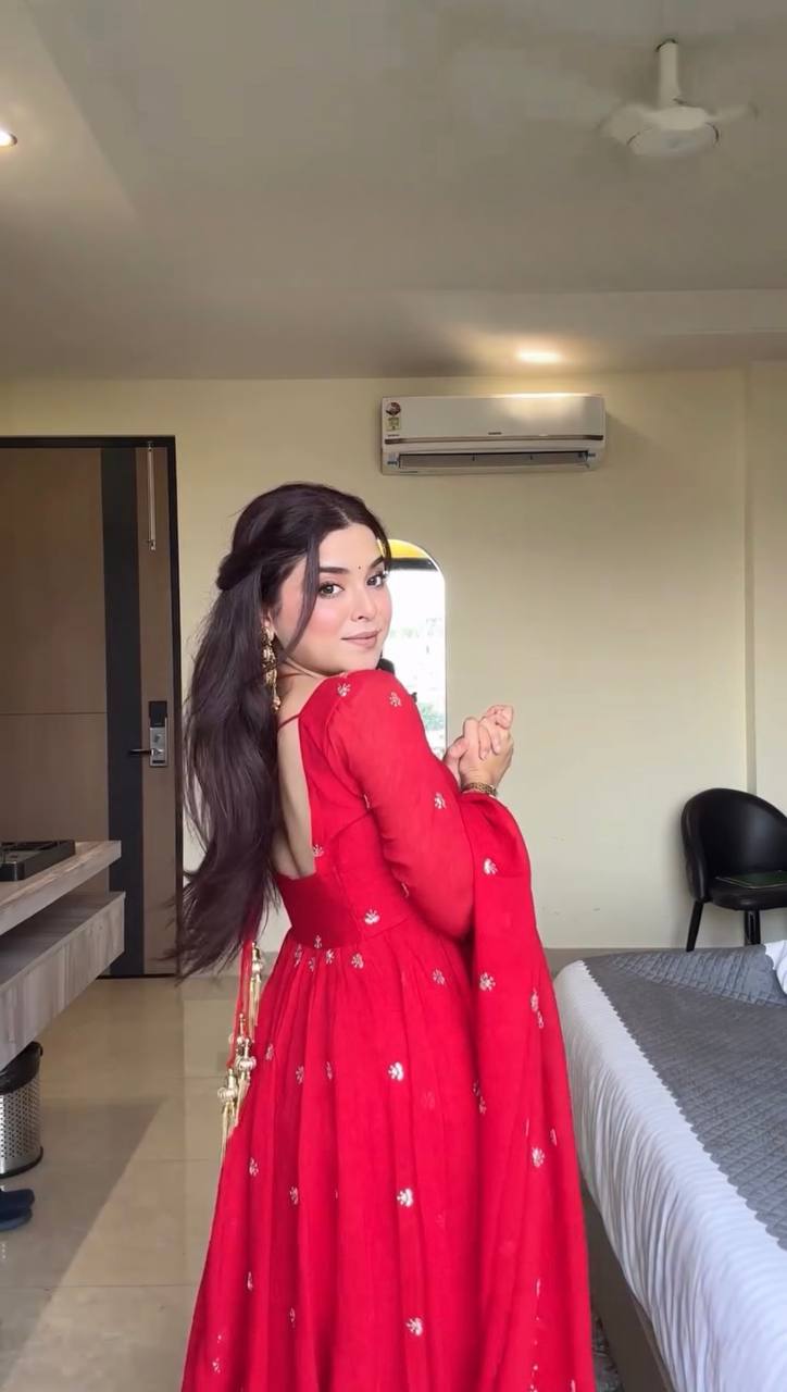 Woman in a red traditional outfit standing in a room with a bed and air conditioner.