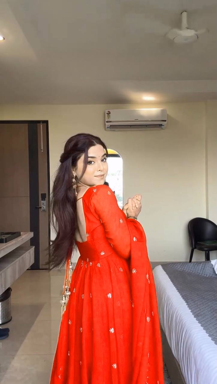Woman in a red traditional outfit standing in a room with a bed and air conditioner.